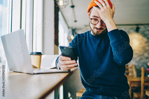 Emotional hipster guy disappointed with banking balance on smartphone sittin in coworking space, stressed young male student shocked with missing lesson checking schedule on mobile phone organizer.