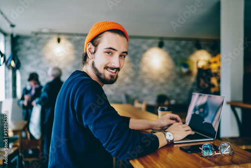 Skilled male it developer looking at camera coding on modern laptop computer working on freelance in cafe interior, portrait of cheerful hipster guy using netbook for e learning satisfied with course.
