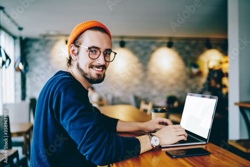 Portrait of happy male freelancer in optical eyewear for vision correction smiling at camera during break from web working online, cheerful hipster blogger sitting in cafe with mockup laptop computer