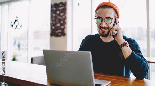 Portrait of cheerful hipster guy talking with friend about time of meeting in coworking space for e learning on laptop computer, happy man connected to public internet for calling via cellphone