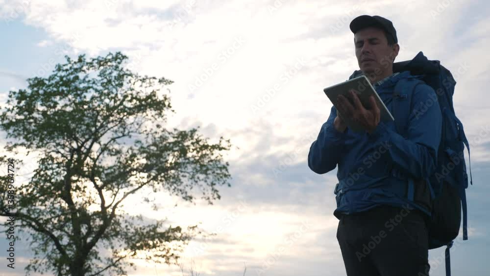 A hipster man with a backpack is traveling, enjoying the landscape, holding a digital tablet. Uses a portable device for navigation, notes, photos, and freelancing. A hipster man with a backpack is traveling, enjoying the landscape, holding a digital tablet. Uses a portable device for navigation, notes, photos, and freelancing.