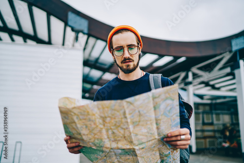 Pensive hipster guy in spectacles reading ma;l while walking on urban setting having trip on vacations, thoughtful caucasian male tourist checking location and destination on route in city tour . photo