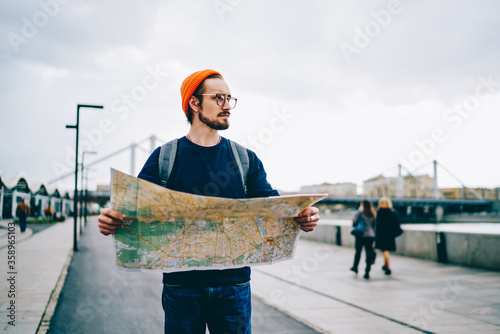 Thoughtful male tourist looking away searching route for destination while walking on urban setting, pensive serious caucasian traveler using map while strolling and checking location on tour