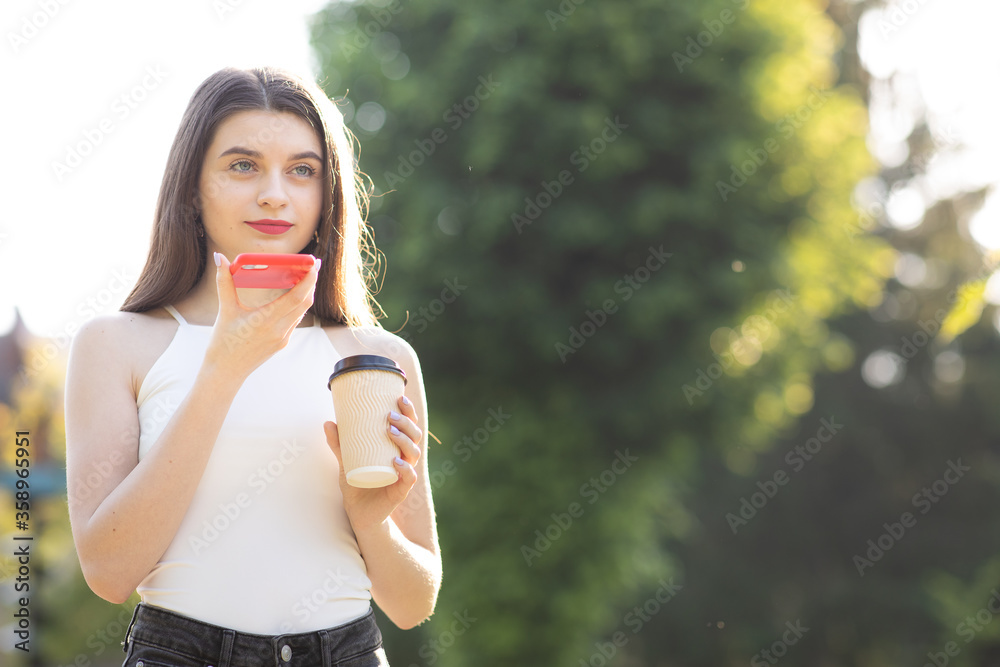 Girl walking in the Park speaking on Smartphone. Happy girl using a smart phone voice recognition audio ai message speech function on line walking in park. Having cup with Coffee in her Hands