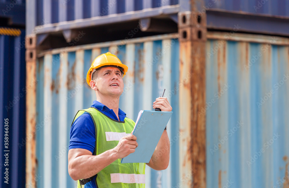 Engineer man in hard hat and safety vest holding clipboard checklist and talks on two-way radio at containers cargo