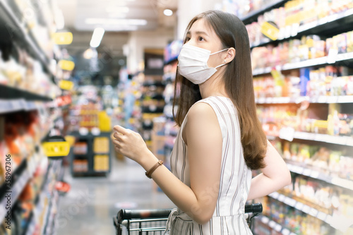 Asian woman wearing face mask push shopping cart in suppermarket . Girl choosing, looking somethings to buy at shelf during coronavirus crisis or covid19 .happy lifestyle or shopaholic concept.