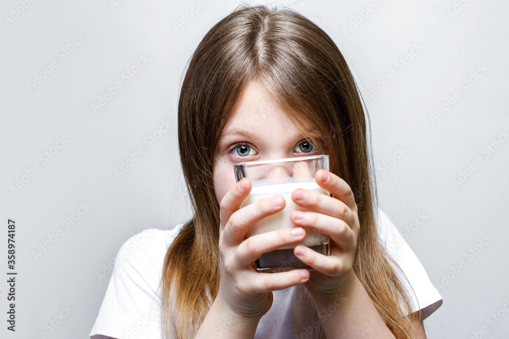 The girl looks out mischievously from behind a glass of milk. Close-up of a pretty girl holding a white milk in a transparent glass. The girl looks out mischievously from behind a glass of milk. Close-up of a pretty girl holding a white milk in a transparent glass.
