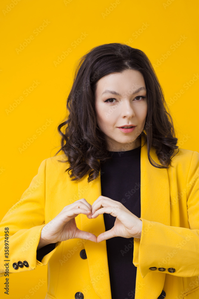 A young brunette woman in a yellow jacket shows a heart gesture. A sign of love and support. A young brunette woman in a yellow jacket shows a heart gesture. A sign of love and support.