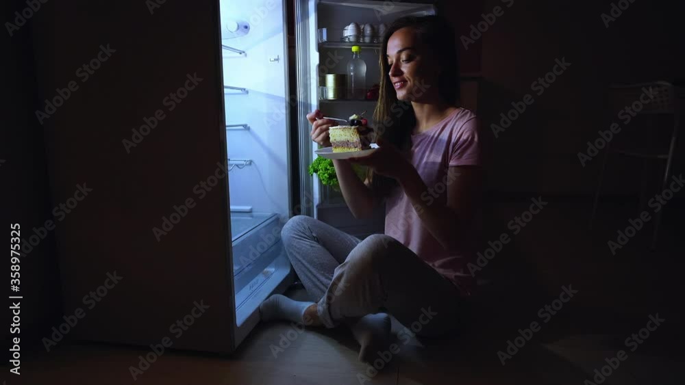 Happy woman eating and enjoying of a sweet tasty piece of cake at night time. Diet fail concept