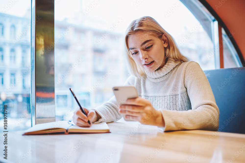 Teen girl reading new message on smartphone while learning in cafe interior prepare homework task,young female hipster checking notification on cellular creating article for blog publication Teen girl reading new message on smartphone while learning in cafe interior prepare homework task,young female hipster checking notification on cellular creating article for blog publication