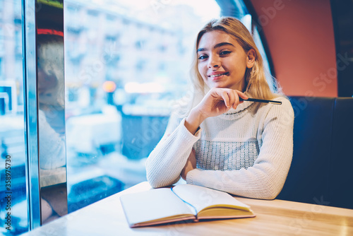 Portrait of cheerful hipster girl satisfied with learning process in cafe interior completing homework, creative female blogger enjoying job and writing texts for publication in notepad on leisure Free Download Portrait of cheerful hipster girl satisfied with learning process in cafe interior completing homework, creative female blogger enjoying job and writing texts for publication in notepad on leisure - GFXLoad