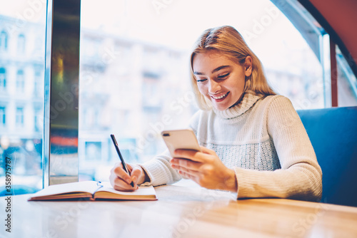 Cheerful female blogger satisfied with getting likes for publication in network holding cellular for reading comments, smiling hipster girl happy checking mail on smartphone 