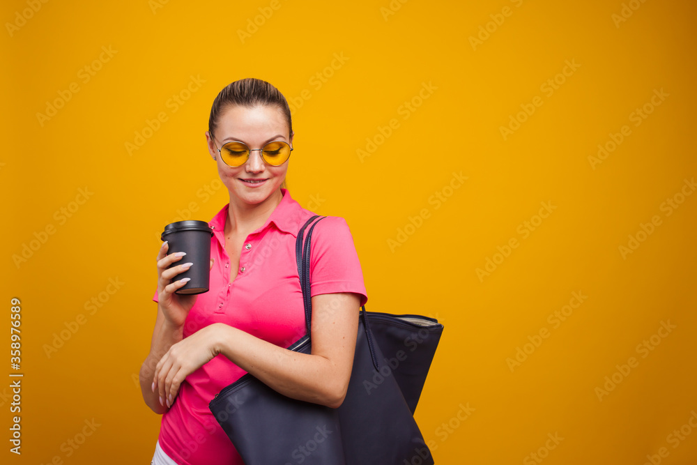 Cute blonde in sunglasses and t-shirt with bag on shoulder, drinking her hot drink, portrait on yellow background