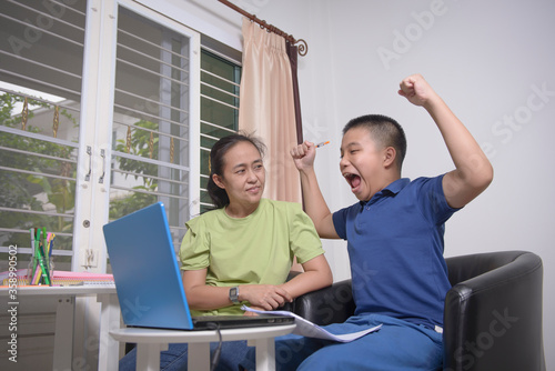 An Asian student appeared very happy when viewing grades on a laptop and he passed the exam in online learning at home. His mother is also congratulating. Concepts of home-learning.