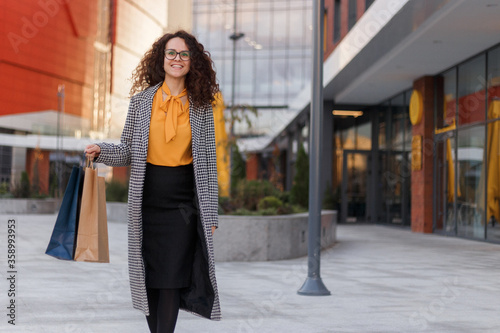 Outdoors portrait of a beautiful smiling curly young woman with many shopping bags, over buildings background. Horizontal view.