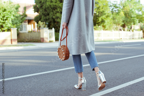 Fashionable young woman wearing grey coat, white blouse, blue jeans, holding straw circle bag with pearls and in silver high heel shoes. Street style.