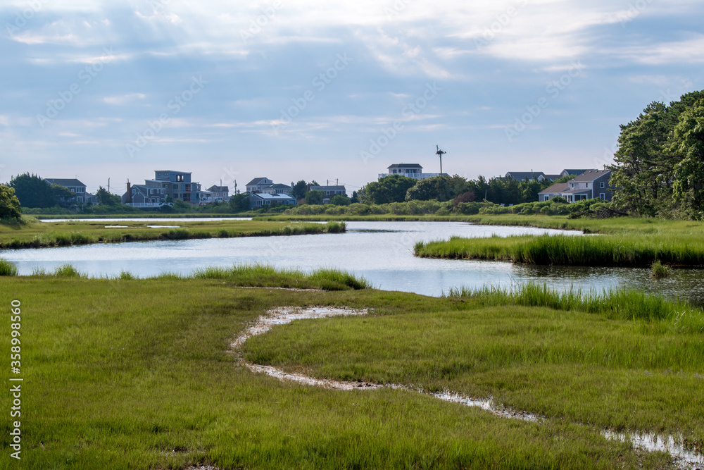 A stream trickling into the calm and serene Bass River in Yarmouth, Cape Cod during summer tourist season as sun rays break through the clouds with an osprey nest in the background.