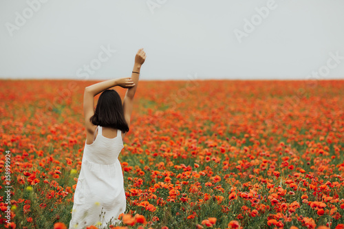 Fototapeta Naklejka Na Ścianę i Meble -  Girl wearing a white dress standing in the middle of a field full of red poppy. Young woman standing with hands up on poppy field from back. Feels happiness, calm and summer vacation. Copy space.