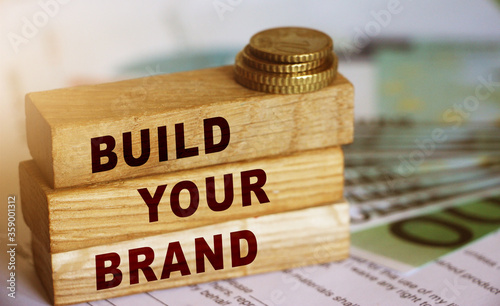 Wooden blocks form the text 'build your brend' on 100 Euro currency bills and coins background. Branding and marketing business concept