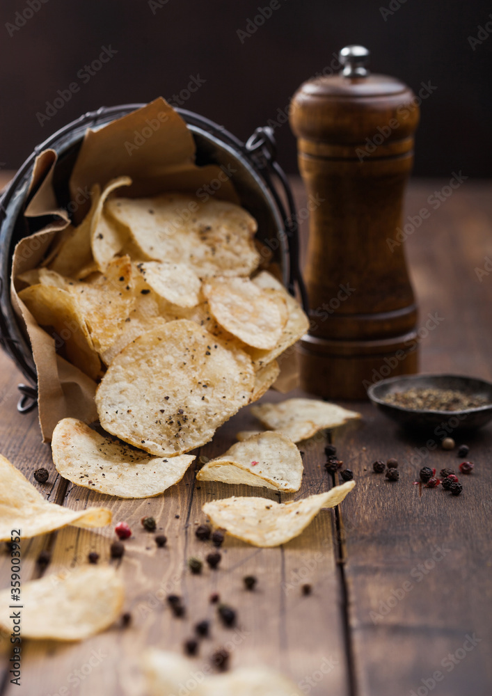 Fototapeta premium Potato crisps chips snack with black pepper in steel bucket on wooden background with mill and ground pepper.