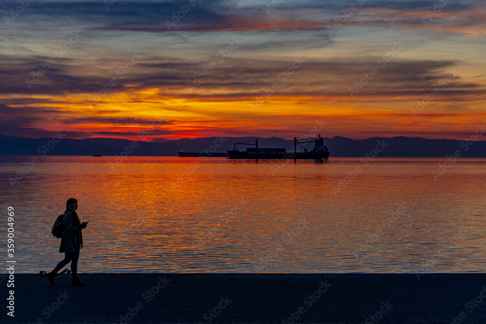 Woman walking at the seafront of Thessaloniki during sunset. Orange and red sky while a container ship lies in the background