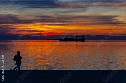 Woman walking at the seafront of Thessaloniki during sunset. Orange and red sky while a container ship lies in the background