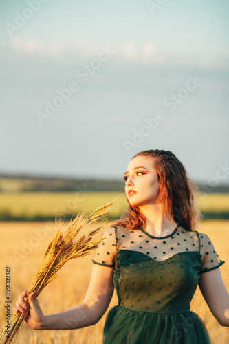 Beautiful woman in green dress on field with sheaf of wheat in hand at sunset light, girl enjoying summer nature landscape, harvest