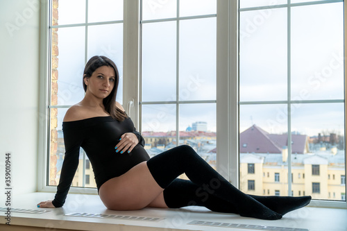 A pregnant woman in the room is sitting on the windowsill, gently putting her hand on her tummy.