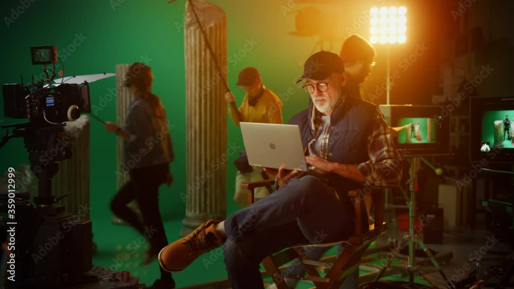 Prominent Successful Director Sitting in a Chair on a Break Using Laptop Computer. On the Studio Film Set with High-End Equipment Professional Crew Shooting High Budget Movie 