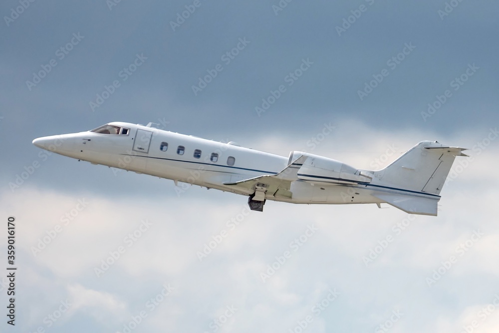 Fototapeta premium Side view of business jet airplane after taking off on grey white clouds in blue sky background. Fast modern aircraft for air transportation. Aviation technology. Travel and business concept.