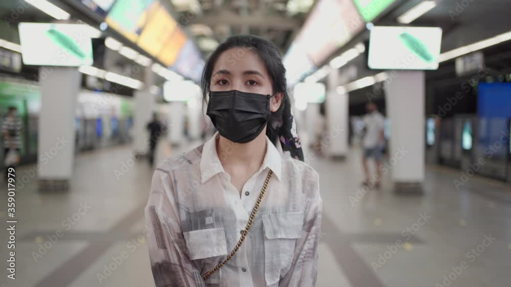 Asian female wear black mask standing and looking at camera in sky train station, covid, woman inside subway station, New normal lifestyle, self protection, public transportation, evening white light Asian female wear black mask standing and looking at camera in sky train station, covid, woman inside subway station, New normal lifestyle, self protection, public transportation, evening white light