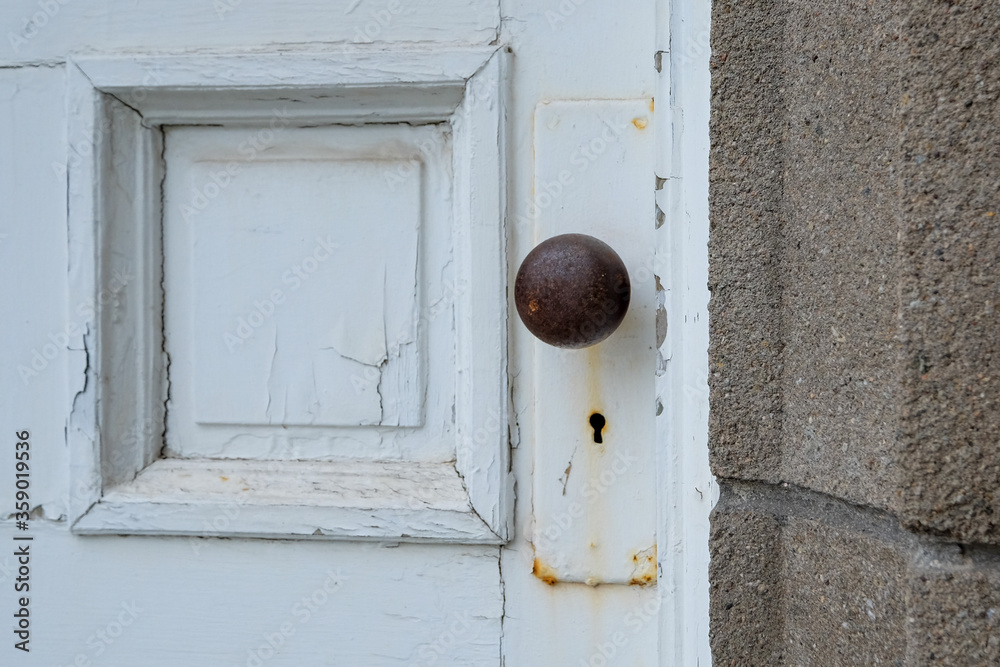 White painted vintage door with a focus on the door knob and keyhole. There's rust on the metal door fixture. A brick wall is adjacent to the door. The knob is a dark brown metal