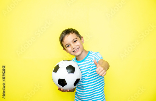 Portrait of emotional toddler boy holding a ball in his hand. Boy in a striped T-shirt on a yellow background. Place for text. Looking at camera