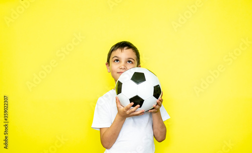 Portrait of an emotional child A boy holds a ball in his hand. A child in a white T-shirt with a ball in his hands looks up on a yellow background. Place for text