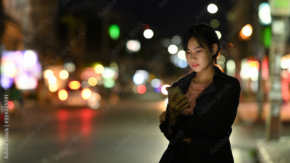 Fototapeta premium Asian woman waits for her private taxi by using a transportation app on the night street.