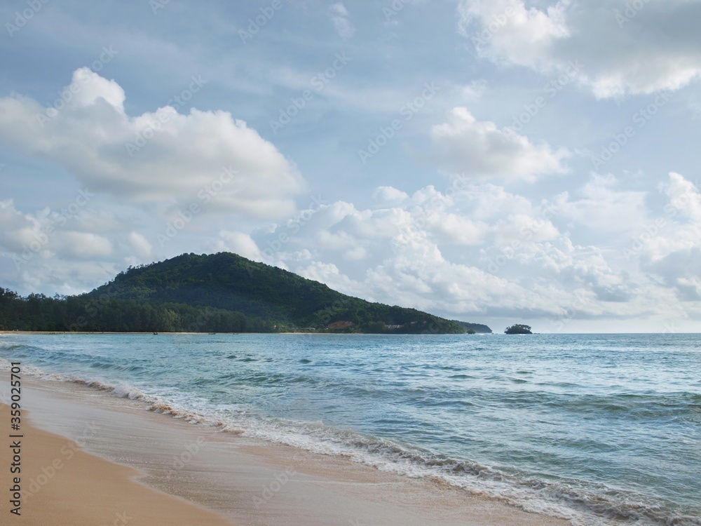 Tropical beach with blue sky and island in Thailand. Magnificent seascape, panoramic view. Mountains, sea, sky, clouds. A green hill covered with dense forest on a horizon. Surf, waves, sand, coast. 