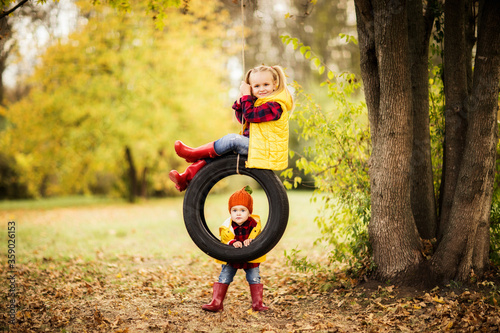 two cute blonde sisters in red rubber boots ride a tire on in autumn Park