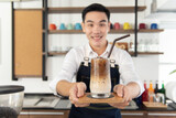 Smiling Asian barista man holding cup of ice beverage to show the  customers in the coffee shop.