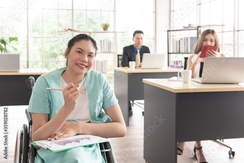 Smiling Asian woman office worker in wheelchair holding pencil look at camera, disabled people working with colleague at workplace