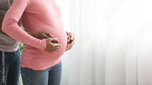 Closeup of african pregnant couple hug, standing next to window