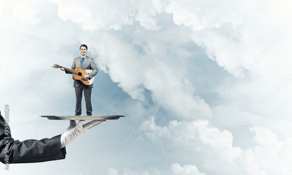 Businessman on metal tray playing acoustic guitar against blue sky background Businessman on metal tray playing acoustic guitar against blue sky background