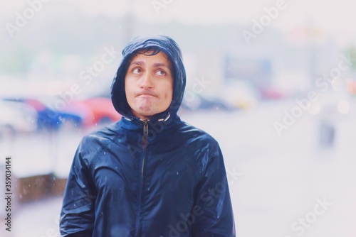 Foto Portrait of a young funny wet man with unhappy tired face in hood walks along the street road during heavy asian rain, close-up