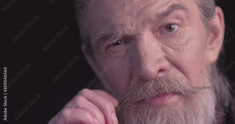 Close up. Old man twists his mustache in black business clothes looking at camera in semi-lit room on black. Toned footage. Prores 422. Close up. Old man twists his mustache in black business clothes looking at camera in semi-lit room on black. Toned footage. Prores 422.