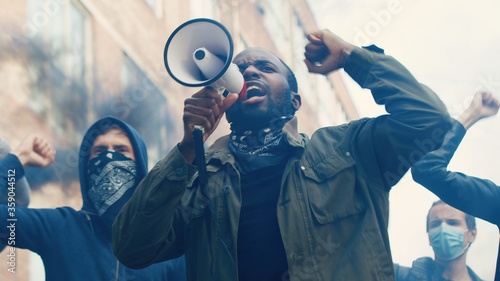 Fototapeta Naklejka Na Ścianę i Meble -  African American young handsome guy with scarf on face protesting in middle of crowd of protesters and screaming mottos in megaphone. Guy leading at manifestation and riot for human rights in smoke.