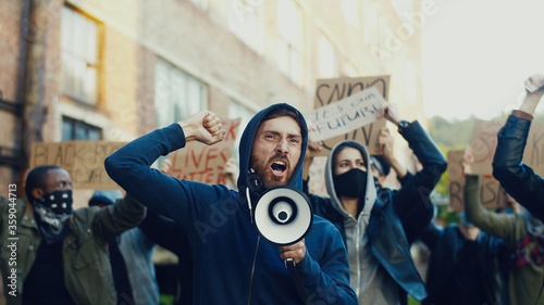 Caucasian young handsome guy protesting in middle of multiethnic crowd of protesters and screaming mottos in megaphone. Guy leading at manifestation for human rights.