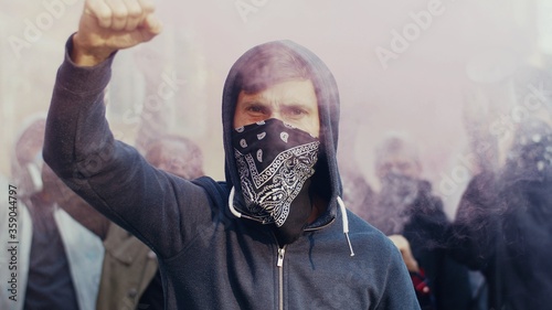 Obraz na plátně Close up of Caucasian handsome young man in hood and scarf on face protesting and screaming slogans in smoke and gas outside