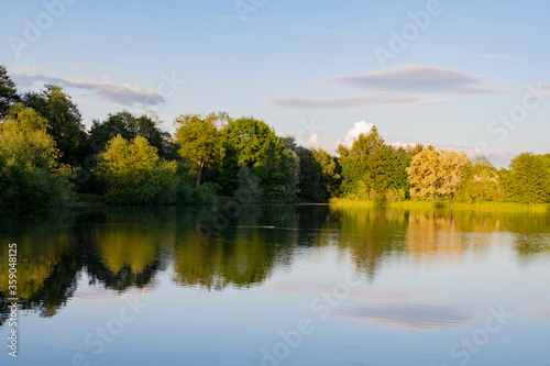 Countryside pond, hills, meadows and forest. Trees and clouds reflecting into the water. Mirror. Warm sunset light. Country pond. Fishing. Countryside holiday