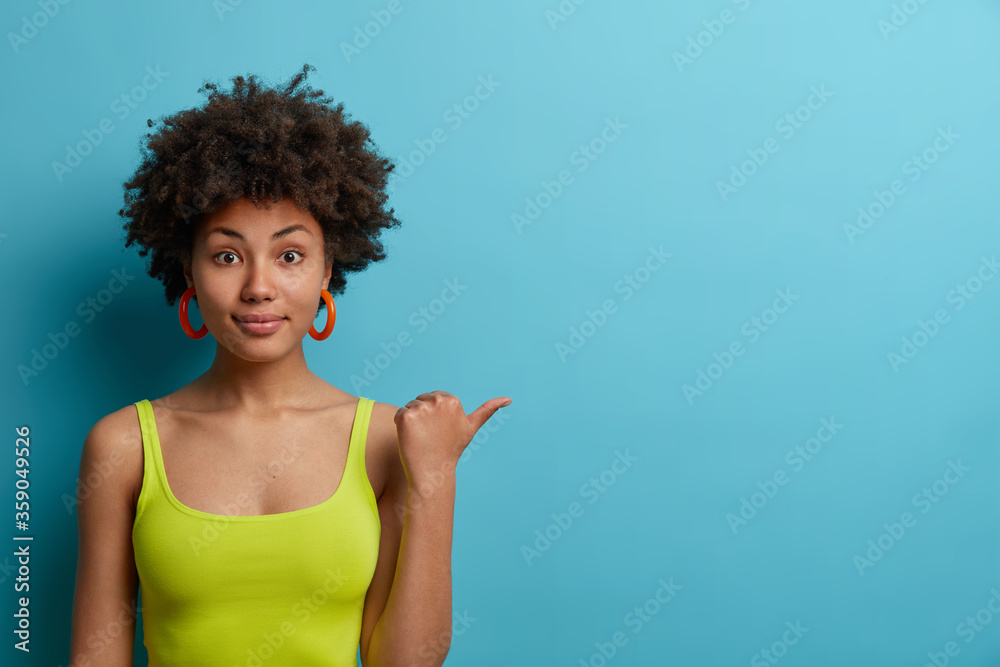 Horizontal shot of calm curly haired Afro American woman points thumb away, wears casual green vest, shows space for your advertisement, isolated on blue background. People, advertisement concept