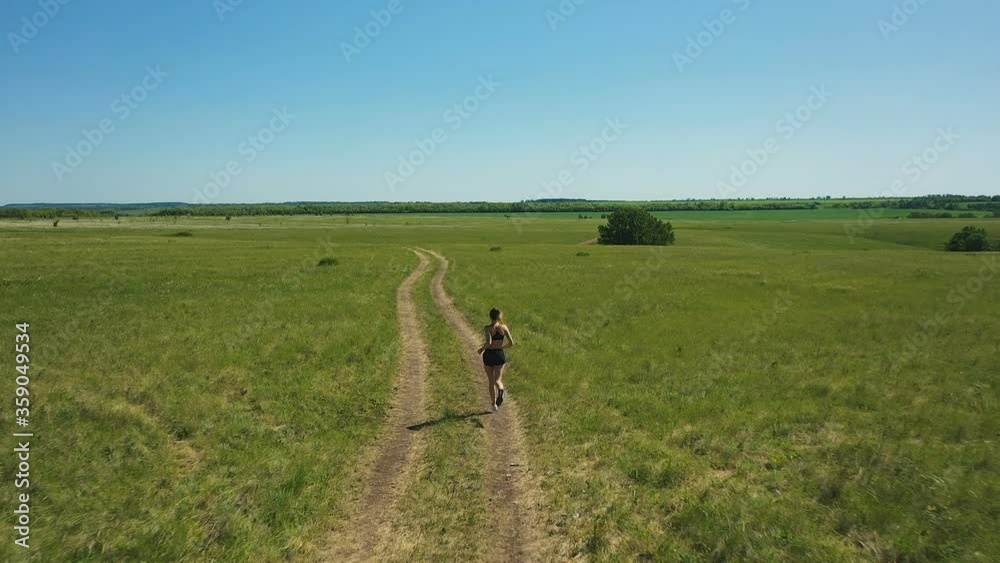 Unrecognizable sportswoman running through field. Aerial tracking shot of slim female in sportswear running along path through green meadow on cloudless day in nature Unrecognizable sportswoman running through field. Aerial tracking shot of slim female in sportswear running along path through green meadow on cloudless day in nature
