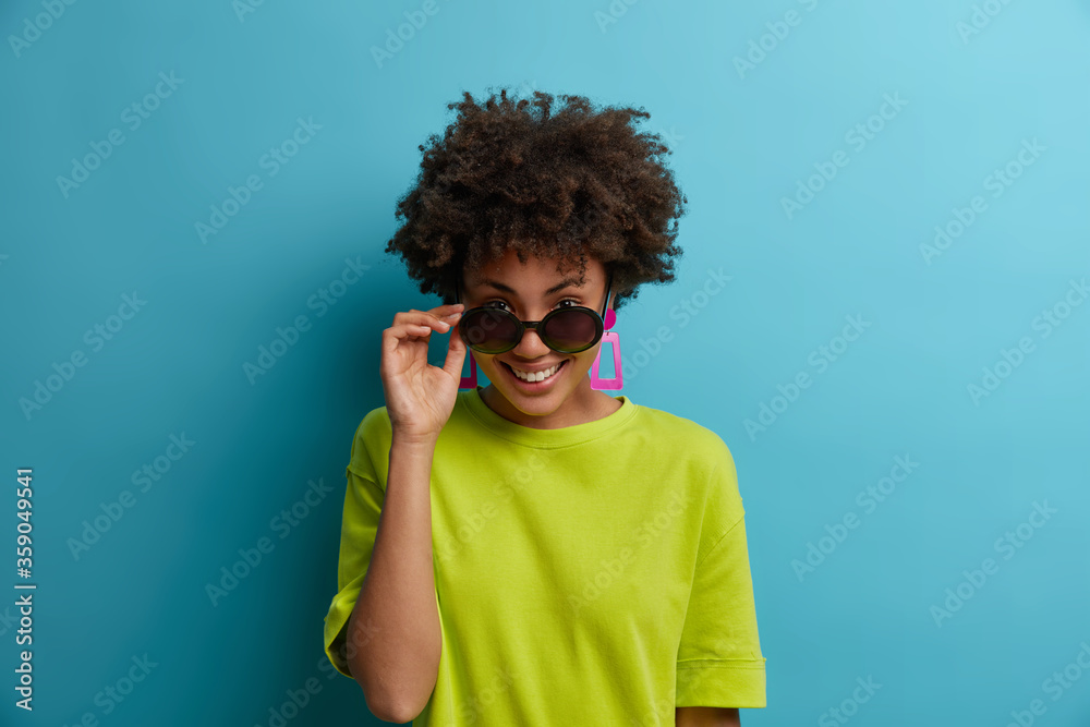 Positive energetic female looks from under sunglasses, enjoys sunny summer day, dressed in casual green t shirt, smiles pleasantly, isolated on blue background. People, season, style concept Positive energetic female looks from under sunglasses, enjoys sunny summer day, dressed in casual green t shirt, smiles pleasantly, isolated on blue background. People, season, style concept